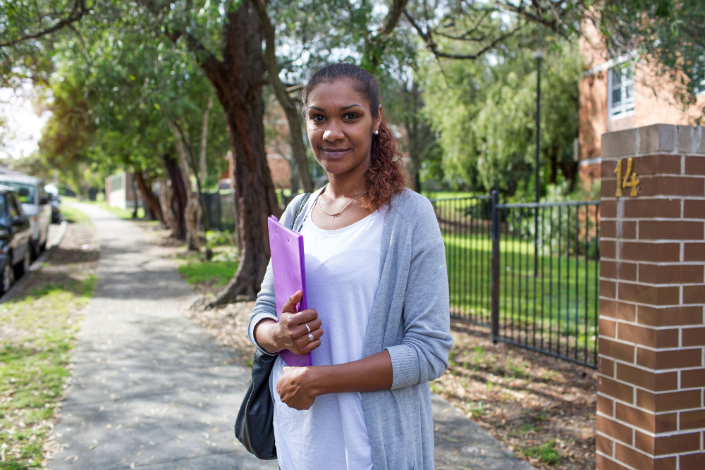 A woman stands in front of an apartment building. She is wearing a white t-shirt and a grey sweater, and is holding a purple folder in her arms.